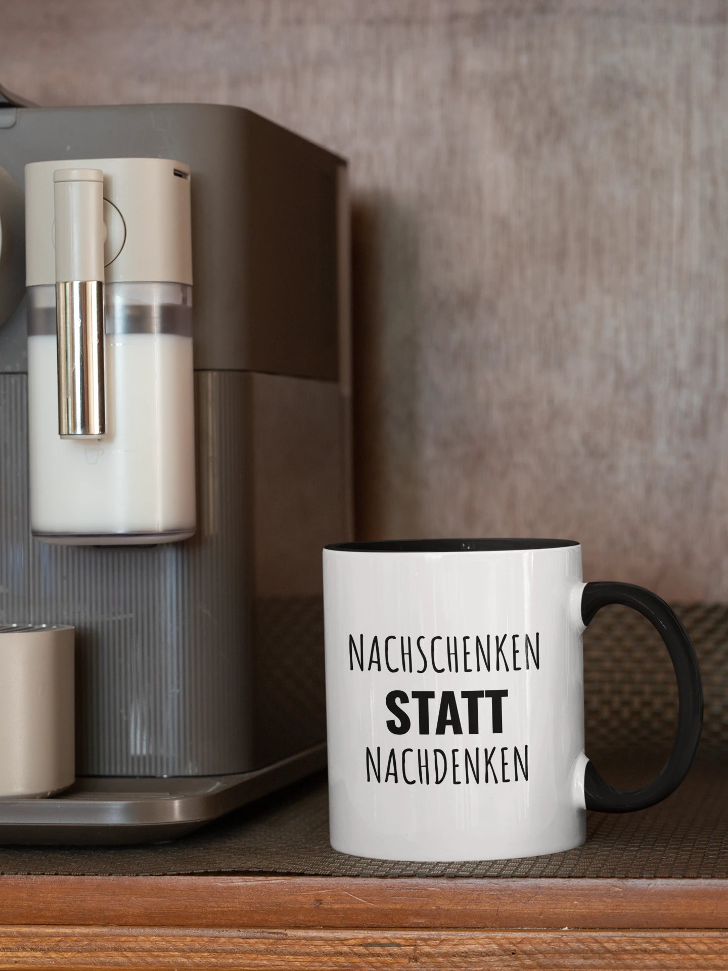 Zweifarbige Tasse mit schwarzem Griff und schwarzer Innenseite, Aufschrift „Nachschenken statt Nachdenken", neben einer Kaffeemaschine